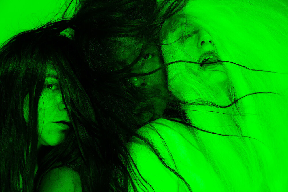Three intertwined bodies and faces, partly veiled by hair, immersed in vivid green.